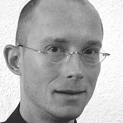 Schwarz-weißes Porträt von Produktdesigner Hammer mit Brille und ruhigem Blick, vor heller Wandstruktur
