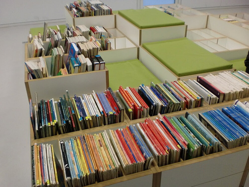 Ein Tojo Regalsystem mit ordentlich sortierten Büchern in verschiedenen Farben und praktischen Aufbewahrungsboxen mit grünen Kissen für zusätzlichen Stauraum und Sitzmöglichkeiten.