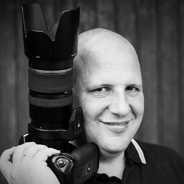 Schwarz-weißes Porträt von Produktdesigner Seegis mit Kamera auf der Schulter, lächelnd vor hölzernem Hintergrund