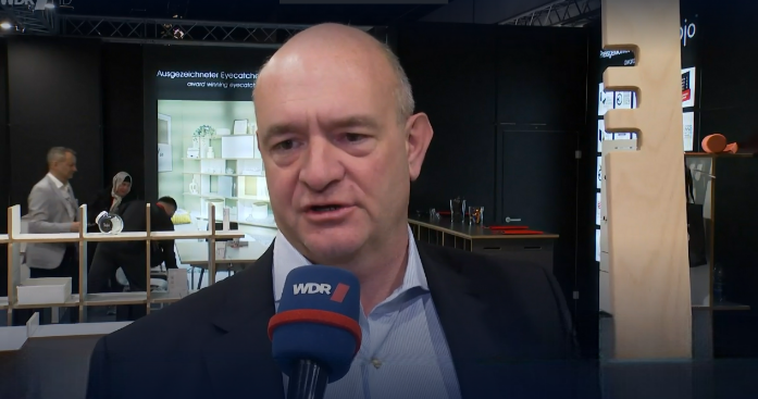 Interviewaufnahme mit Tojo-Vertreter vor Messestand, spricht ins WDR-Mikrofon – im Hintergrund moderne Möbel und Gesprächssituation