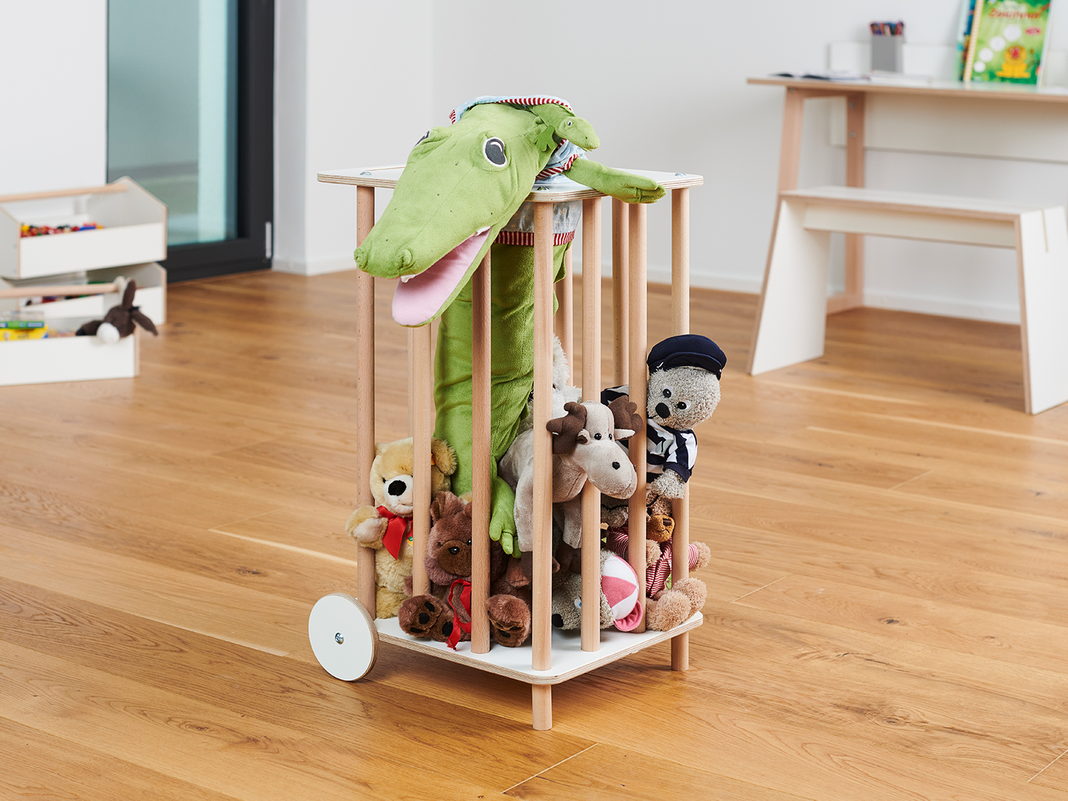 Mobiler Spielzeugkorb Tojo-zoo mit Holzstäben und weißen Rollen, gefüllt mit Kuscheltieren in einem hellen Kinderzimmer mit Holzfußboden