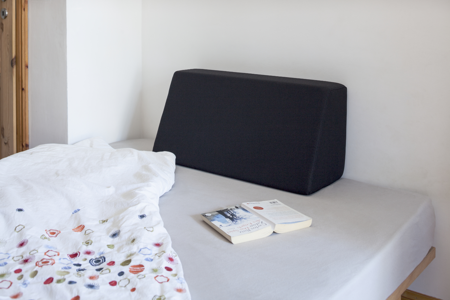 Tojo-Lehne in Schwarz auf einem schlichten Bett platziert, mit aufgeschlagenem Buch und heller Bettwäsche – gemütliches Setting für Lesen oder Entspannen