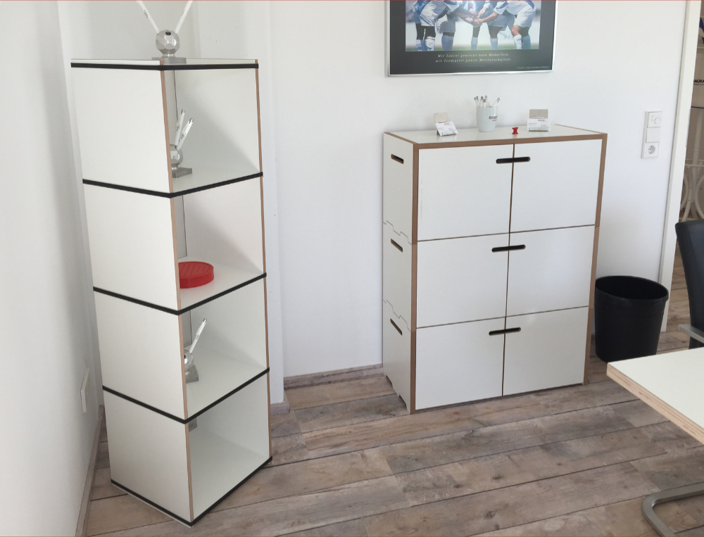 Zwei Tojo Aufbewahrungseinheiten im Büro, eine schmale Regalstruktur und ein größerer Schrank, beide in weiß mit markanten roten Akzenten an den Türen, bieten funktionalen Stauraum und einen modernen Look.