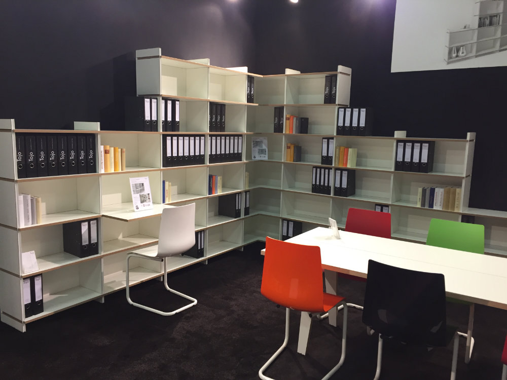 Tojo Regal „mehrfach“ als große Regalwand in Messeumgebung mit schwarzer Rückwand und bunten Stühlen – vielseitig bestückt mit Ordnern und Büchern, ideal für Büro oder Ausstellung
