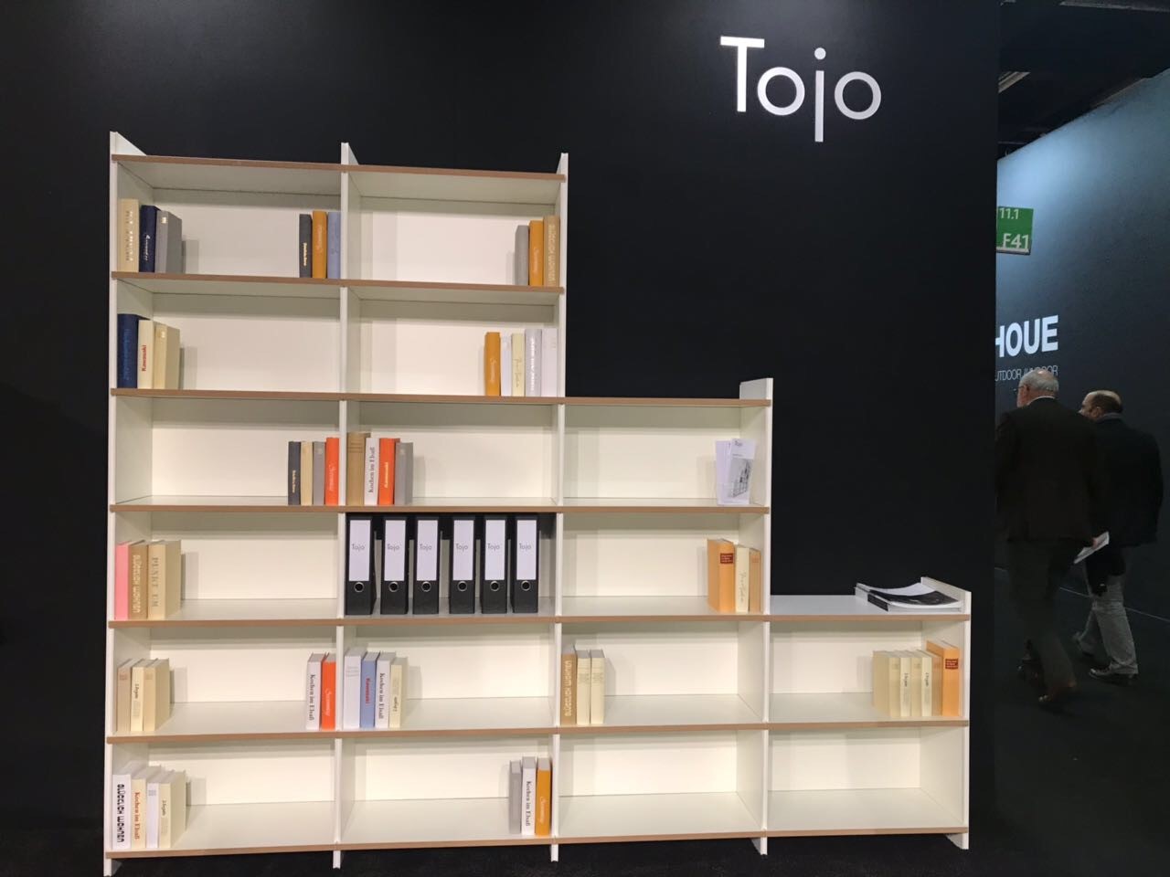 Stufenförmig angeordnetes Regal „mehrfach“ von Tojo in Weiß mit naturbelassenem Holzrahmen, präsentiert auf einer Messewand – offenes Regalsystem für Bücher, Ordner und Dekoration