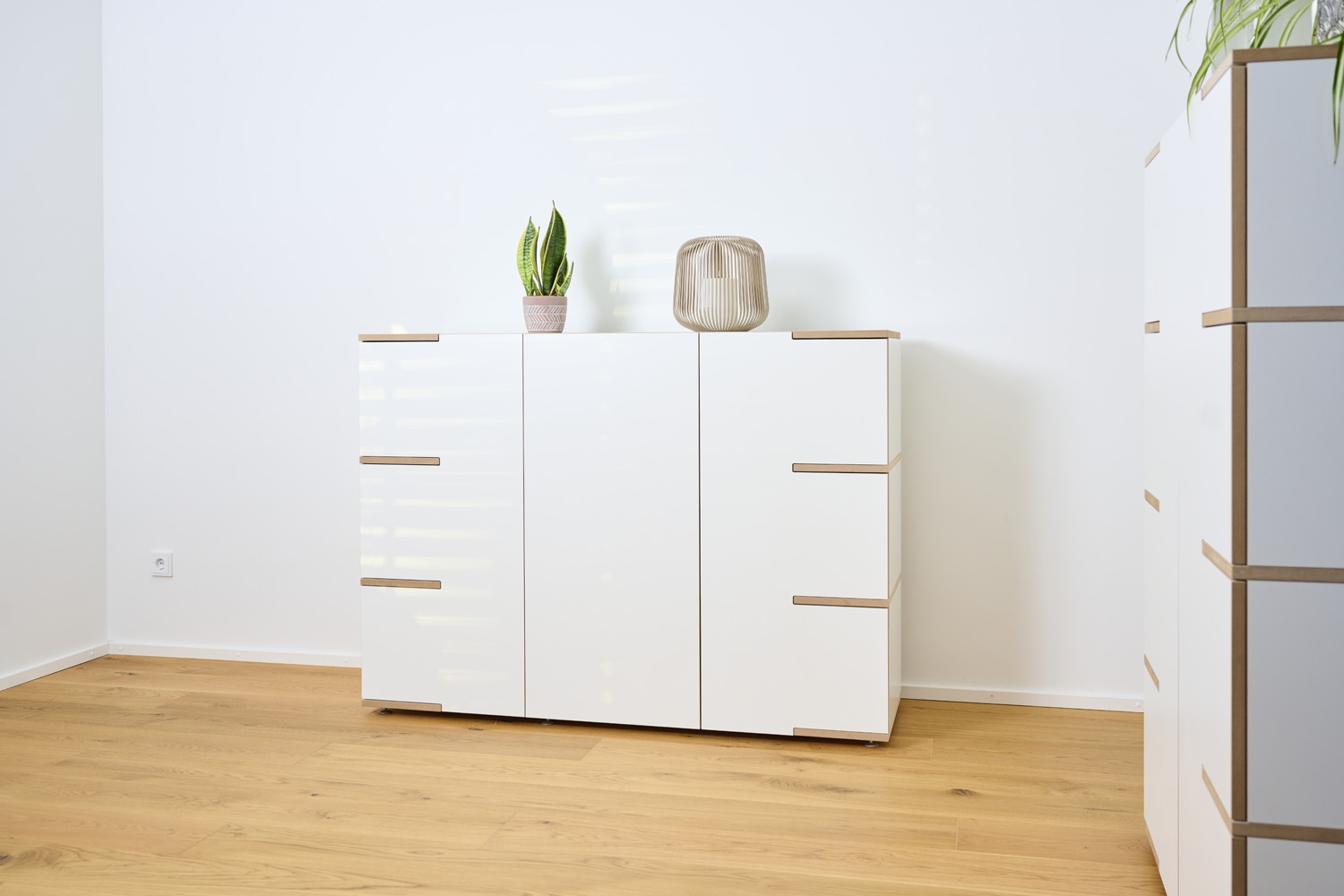 Weißes Sideboard Tojo stau mit drei Schrankmodulen, dekorativem Aufbau und Vasen auf der Oberfläche, platziert in einer hellen Raumecke