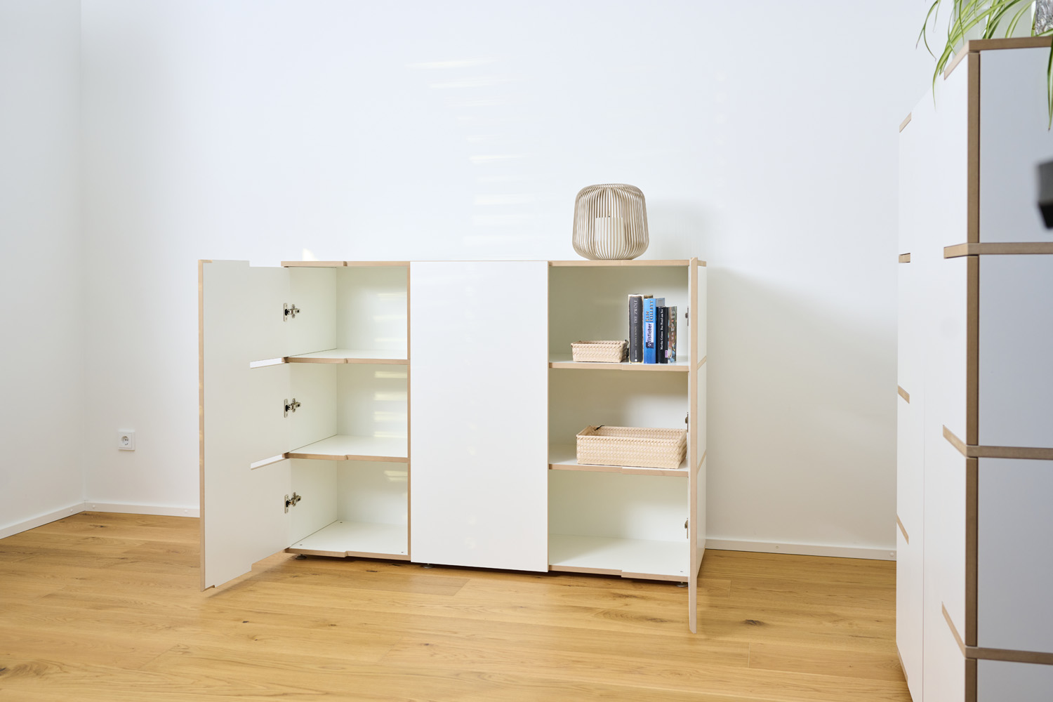 Geöffnetes weißes Sideboard Tojo stau mit sechs geräumigen Fächern, teilweise mit Büchern und Deko befüllt, stehend in einer hellen Raumecke