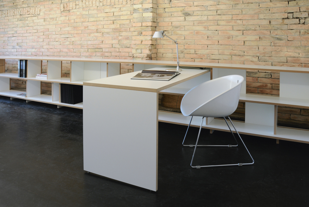 Modularer Schreibtisch Tojo anstell mit weißer Arbeitsfläche und integrierter Regalwand vor einer Ziegelwand in einem modernen Büro