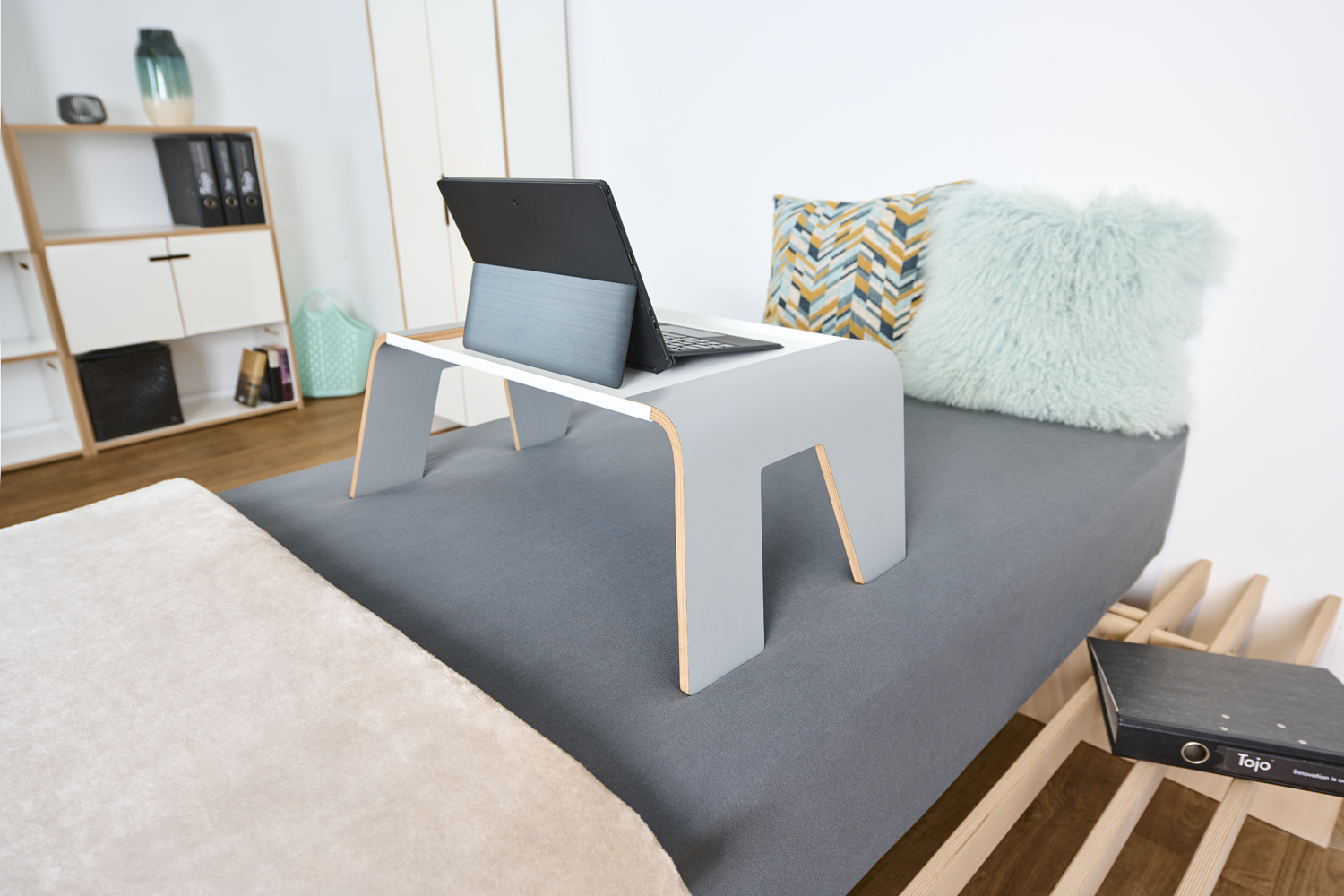 Der Tojo praktisch Tisch steht auf einem Bett, dient als Ablage für einen Laptop und ist von Kissen umgeben